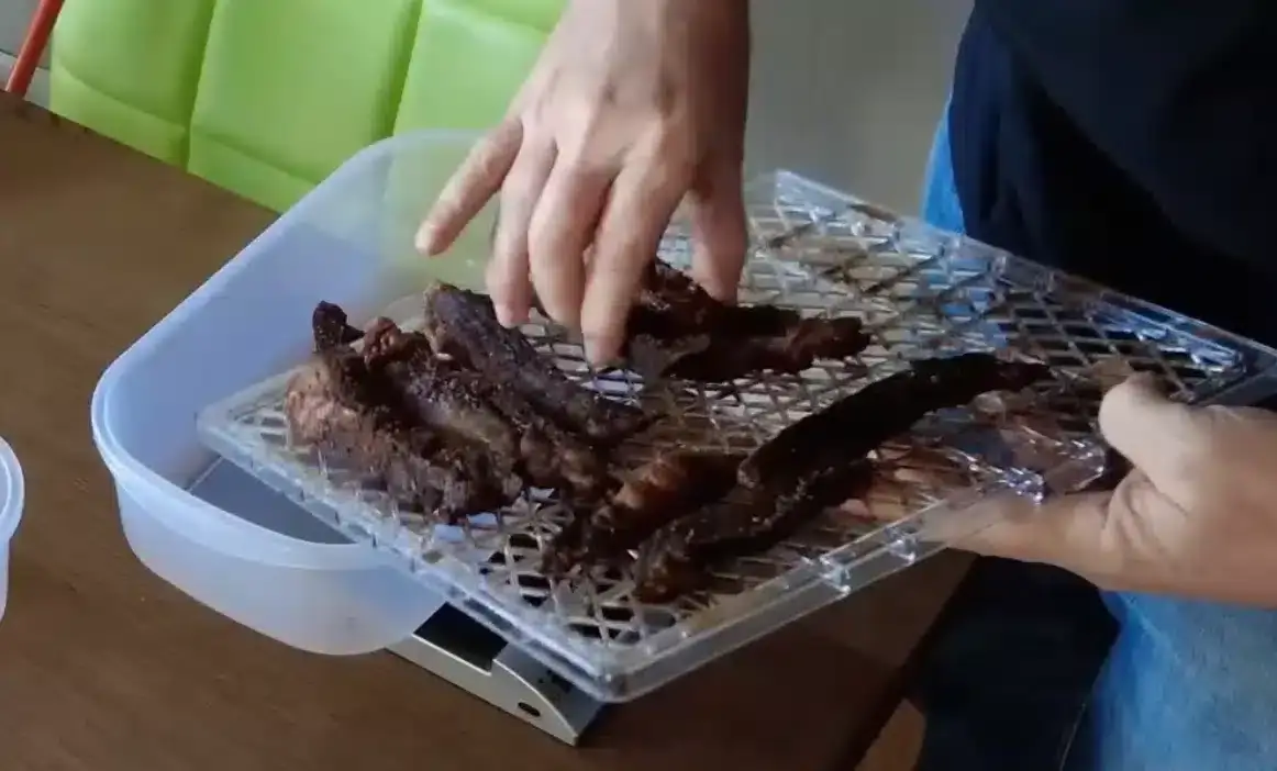 A person placing homemade biltong from a drying rack into a plastic container. Perfect for a DIY biltong guide.